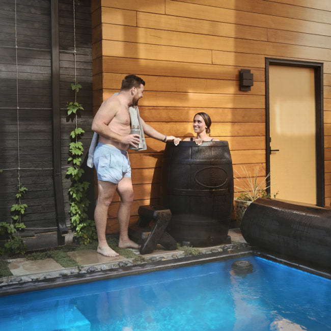 An athletic man stands outside next to his wife whom is enjoying an ice bath