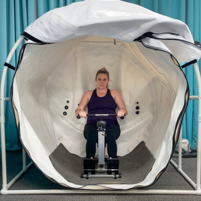 An interior view of the large Summit To Sea Grand Dive Pro Plus Hyperbaric chamber. A woman sits inside exercising on a rowing machine